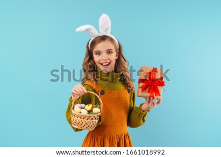 Photo of joyful blonde girl in toy rabbit ears holding basket with eggs and gift box isolated over blue background