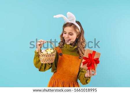 Photo of joyful blonde girl in toy rabbit ears holding basket with eggs and gift box isolated over blue background