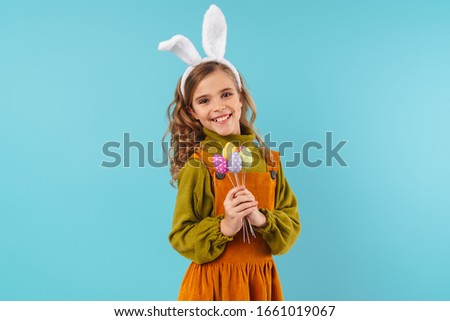 Photo of joyful blonde girl in toy rabbit ears smiling and holding colorful eggs isolated over blue background