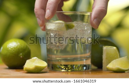Close up hand shaking a glass of soda water. Tonic fizzy water with ice and piece of lime on a green background. Refreshing mineral water, preparing of drinks, cocktails. Cold lemonade drink