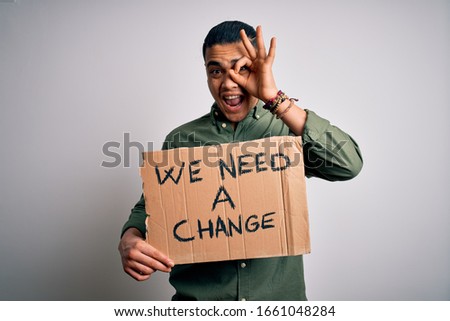 Young brazilian activist man asking for change holding banner over white background with happy face smiling doing ok sign with hand on eye looking through fingers