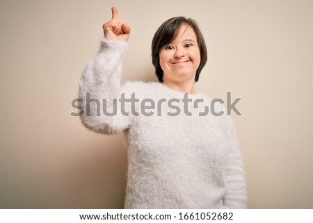 Young down syndrome woman standing over isolated background showing and pointing up with finger number one while smiling confident and happy.