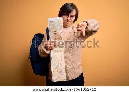 Young down syndrome tourist woman wearing bag and looking at destination city map with angry face, negative sign showing dislike with thumbs down, rejection concept