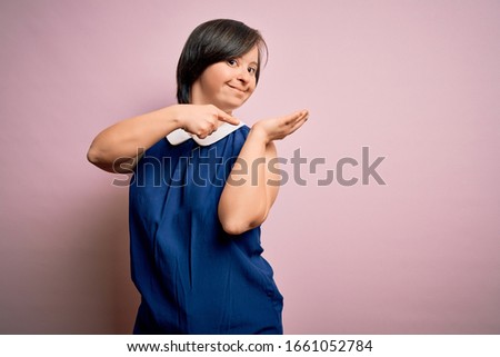 Young down syndrome woman wearing elegant shirt over pink background amazed and smiling to the camera while presenting with hand and pointing with finger.