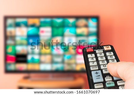 Caucasian man choosing what to watch on TV at home. Video on demand or VOD abstract concept. Changing channels and adjusting volume with television remote control.  Royalty-Free Stock Photo #1661070697