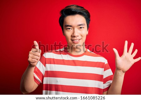 Young handsome chinese man wearing casual striped t-shirt standing over red background showing and pointing up with fingers number six while smiling confident and happy.