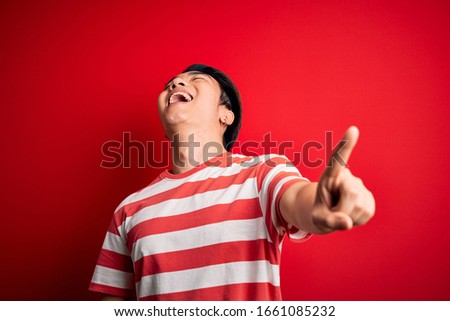 Young handsome chinese man wearing casual striped t-shirt standing over red background laughing at you, pointing finger to the camera with hand over body, shame expression
