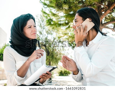 Serious business woman talking on cellphone outside. Her Muslim female colleague in hijab listening to her and writing down notes. Teamwork concept