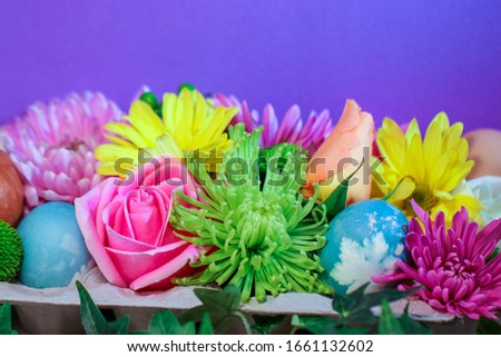 A collection of colorful fresh florist flowers displayed with natural-dyed eggs with botanical leaf prints in an egg carton against a solid purple background, with copy space