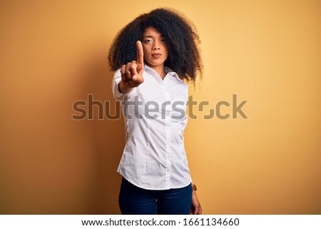Young beautiful african american elegant woman with afro hair standing over yellow background Pointing with finger up and angry expression, showing no gesture