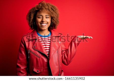 Young beautiful African American afro woman with curly hair wearing casual red jacket smiling cheerful presenting and pointing with palm of hand looking at the camera.