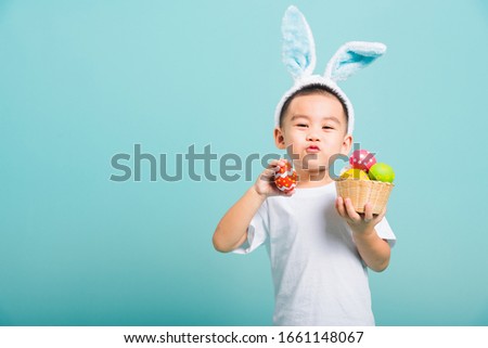 Asian cute little child boy smile beaming wearing bunny ears and a white T-shirt, standing to hold a basket with full Easter eggs. And other hand holds an easter egg on blue background with copy space