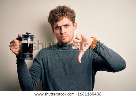 Young blond man with curly hair making coffee using coffemaker over white background with angry face, negative sign showing dislike with thumbs down, rejection concept
