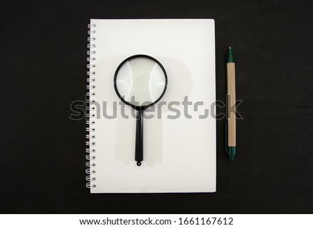 white notebook with magnifier and pen on a black table. View from above.