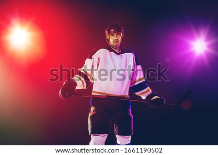 Winner. Male hockey player with the stick on ice court and dark neon colored background. Sportsman wearing equipment, helmet practicing. Concept of sport, healthy lifestyle, motion, wellness, action.