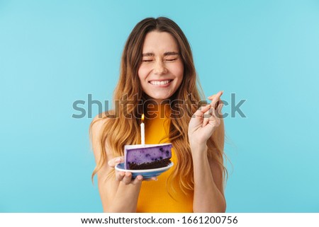 Portrait of joyous woman making wish while holding birthday cake with candle isolated over blue background in studio