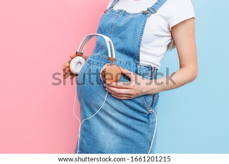 Close - up of a pregnant woman's belly holding headphones with music near her belly, on a pink and blue background