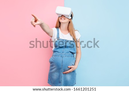 A cheerful happy pregnant woman has fun wearing virtual reality glasses while standing on a pink and blue background