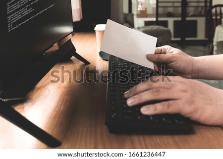 A programmer works in front of desktop computer and picking a piece of paper in right hand 