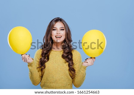 Optimistic stylish girl showing yellow balloons and smiling for camera while having fun against blue background
