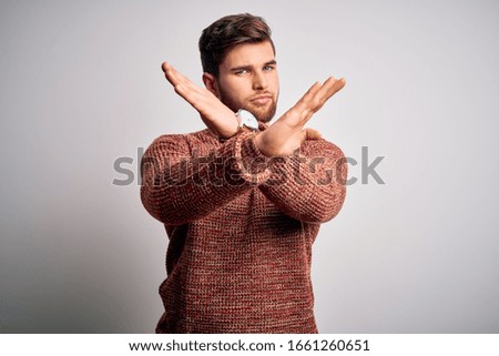Young blond man with beard and blue eyes wearing casual sweater over white background Rejection expression crossing arms and palms doing negative sign, angry face