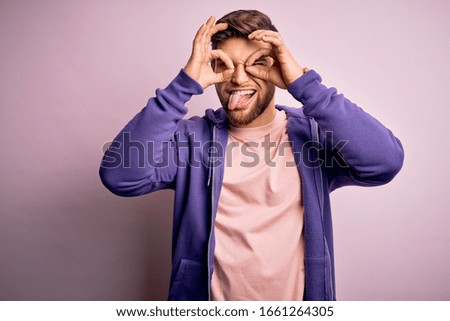 Young blond man with beard and blue eyes wearing purple sweatshirt over pink background doing ok gesture like binoculars sticking tongue out, eyes looking through fingers. Crazy expression.