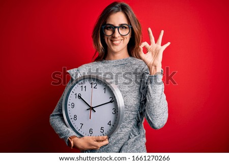Young beautiful brunette woman doing countdown holding big clock over red background doing ok sign with fingers, excellent symbol