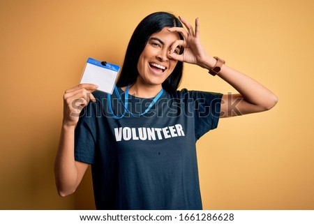 Young beautiful hispanic woman wearing volunteer t-shirt and showing identification with happy face smiling doing ok sign with hand on eye looking through fingers