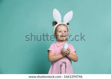 Small blond happy girl in white decorative fur bunny ears standing and holding blue egg in hands on pastel blue background. Family look and celebrating Easter holiday concept
