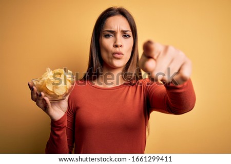 Young beautiful brunette woman holding bowl with chips potatoes over yellow background pointing with finger to the camera and to you, hand sign, positive and confident gesture from the front