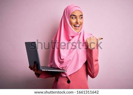 Young beautiful businesswoman wearing pink muslim hijab and business jacket using laptop pointing and showing with thumb up to the side with happy face smiling