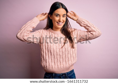 Young beautiful brunette woman wearing casual sweater over isolated pink background Smiling pulling ears with fingers, funny gesture. Audition problem