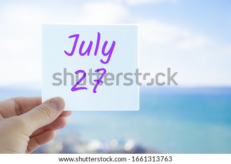 July 27th. Hand holding sticker with text July 27 on the blurred background of the sea and sky. Copy space for text. Month in calendar concept