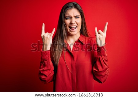 Young beautiful woman with blue eyes wearing casual shirt standing over red background shouting with crazy expression doing rock symbol with hands up. Music star. Heavy concept.
