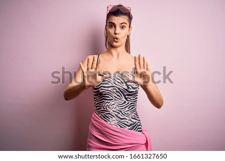 Young beautiful brunette woman on vacation wearing swimsuit over pink background Moving away hands palms showing refusal and denial with afraid and disgusting expression. Stop and forbidden.