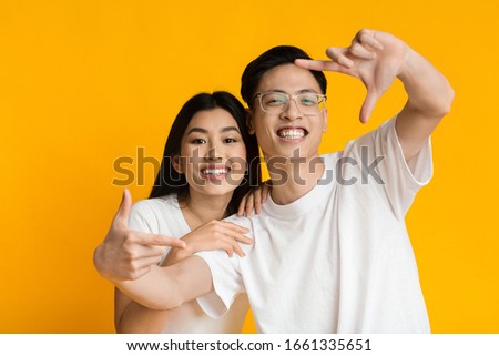 Asian couple laughing and showing frame sign with their fingers on yellow studio background