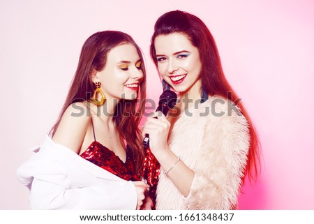 Beautiful brunettes on a pink background sing into a microphone. Teenager in karaoke. Girl with bright green make-up, earrings and red lips. Sisters on a pink background.