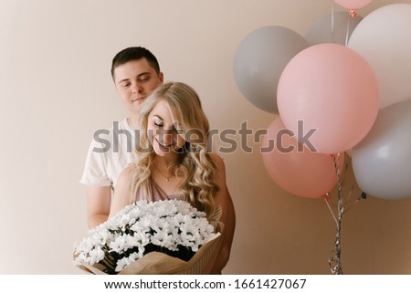 Beautiful young smiling blonde woman and guy with balloons and flowers of white daisies or chrysanthemums on a light background