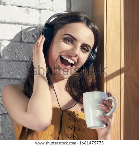 Happy girl listening to music and holding white cup beside window under sunlight