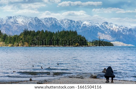 A woman crouching down taking photos of ducks on Lake Wakatipu Queenstown New Zealand with a forest and snow covered alpine mountains in the background