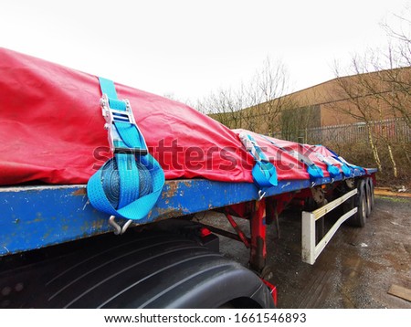 Semi Truck Trailer with a Tarp Covering the Steel Cargo, showing Close up of the Side of the Trailer with Blue Ratchet Straps Coiled up and Securing the Load underneath. Royalty-Free Stock Photo #1661546893