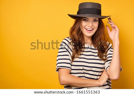 Lovely brunette in a striped T-shirt smiles while holding the edge of a wicker hat on a yellow background.