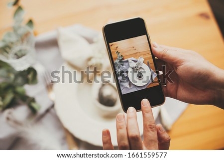 Hand holding phone and taking photo of stylish Easter flat lay table setting with egg in easter bunny napkin. Modern Easter table decorations workshop. Photo for social media