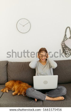 Blonde in a gray blouse and jeans sits on a gray sofa. Girl behind a laptop covers her ears with her hands. Dog is sitting near the girl. String bag and watch on a white wall.