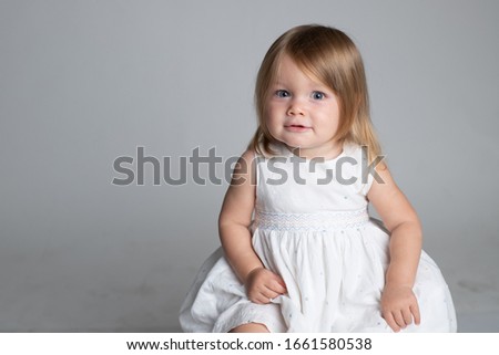 Portrait of a little girl in a white dress. 2 years baby smiling, charmingly looking at the camera. Blonde hair, blue eyes. Sits alone. Hands are visible. Horizontal. Studio lighting, gray background.