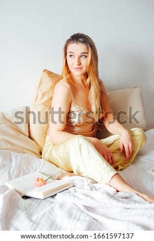 Blonde woman in satin yellow pajamas having breakfast in bed feeling cozy and relaxed