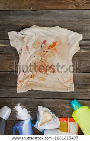 Dirty children's clothes are scattered on a wooden table next to washing powders and soap.Concept washing dirty spots, the best means of cleaning kids clothes.vertically