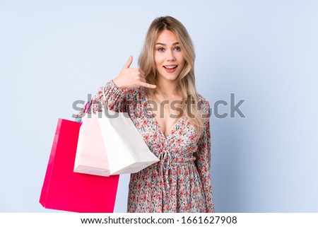 Teenager Russian girl with shopping bag isolated on blue background making phone gesture. Call me back sign