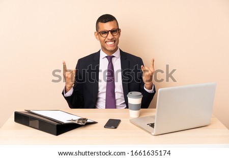 Young business man in his office with a laptop and other documents making rock gesture