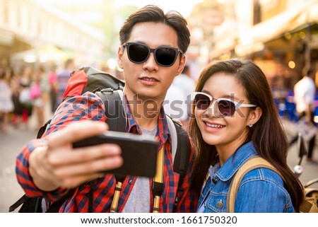 Smiling young Asian couple tourists taking selfie while traveling in Khao San road Bangkok Thailand during summer vacation